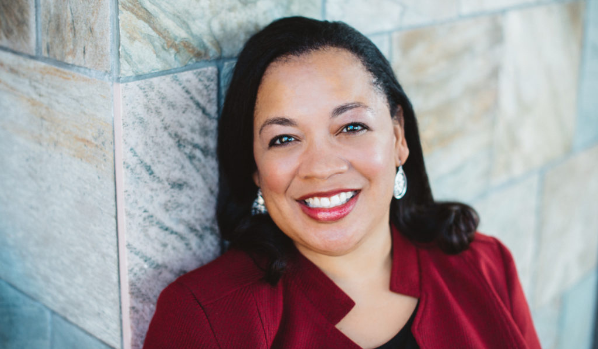 Dr. Saundra Dalton-Smith in a burgundy jacket leans against a textured stone wall, showcasing elegant earrings and long, flowing hair