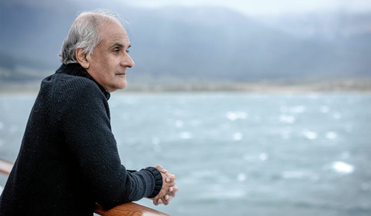 Pico Iyer sits pensively by a lake, overlooking the water with mountains in the background, reflecting a sense of calm solitude