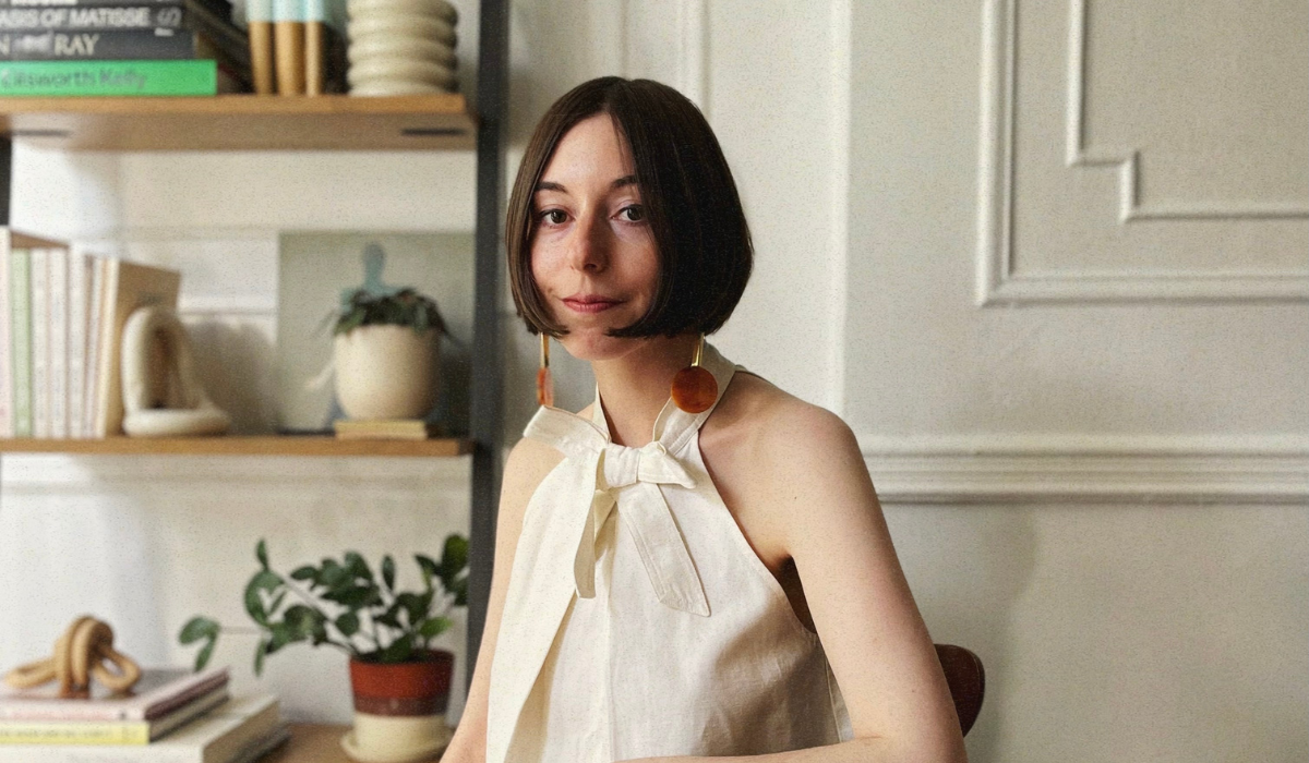 Rachel Schwartzmann in a white halter top sits in a cozy, sunlit room with bookshelves and houseplants in the background