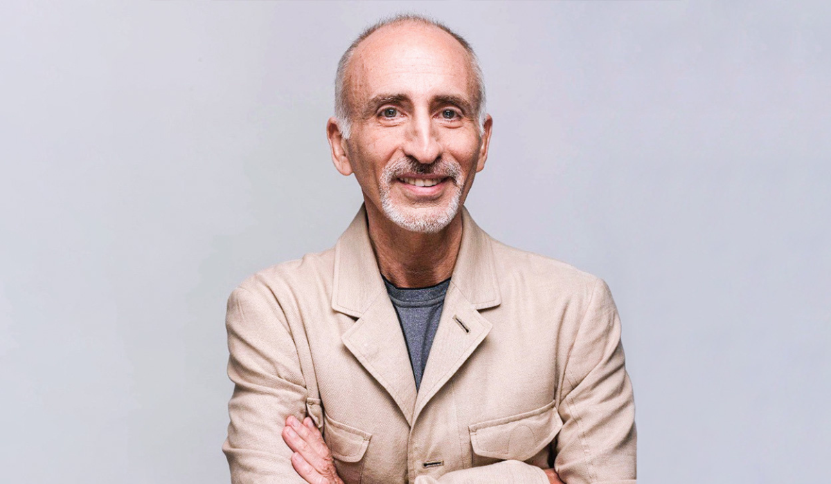 Psychiatrist Phil Stutz wearing a light beige jacket over a dark shirt stands with arms crossed against a plain backdrop