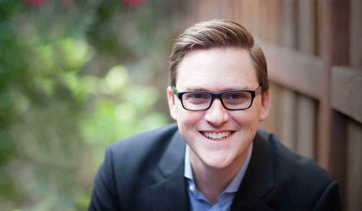 Author Greg McKeown in a dark blazer poses outdoors in front of a blurred greenery background, confident and relaxed