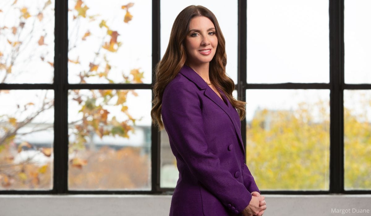 Shannon Watts in a purple blazer stands in profile near large windows with autumn leaves visible outside